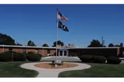 Single-story brick building with a flat roof, surrounded by neatly trimmed bushes and a circular walkway leading to the entrance. Two flagpoles stand in front of the building, one with the American flag and the other with a dark flag. The sky is clear and blue.