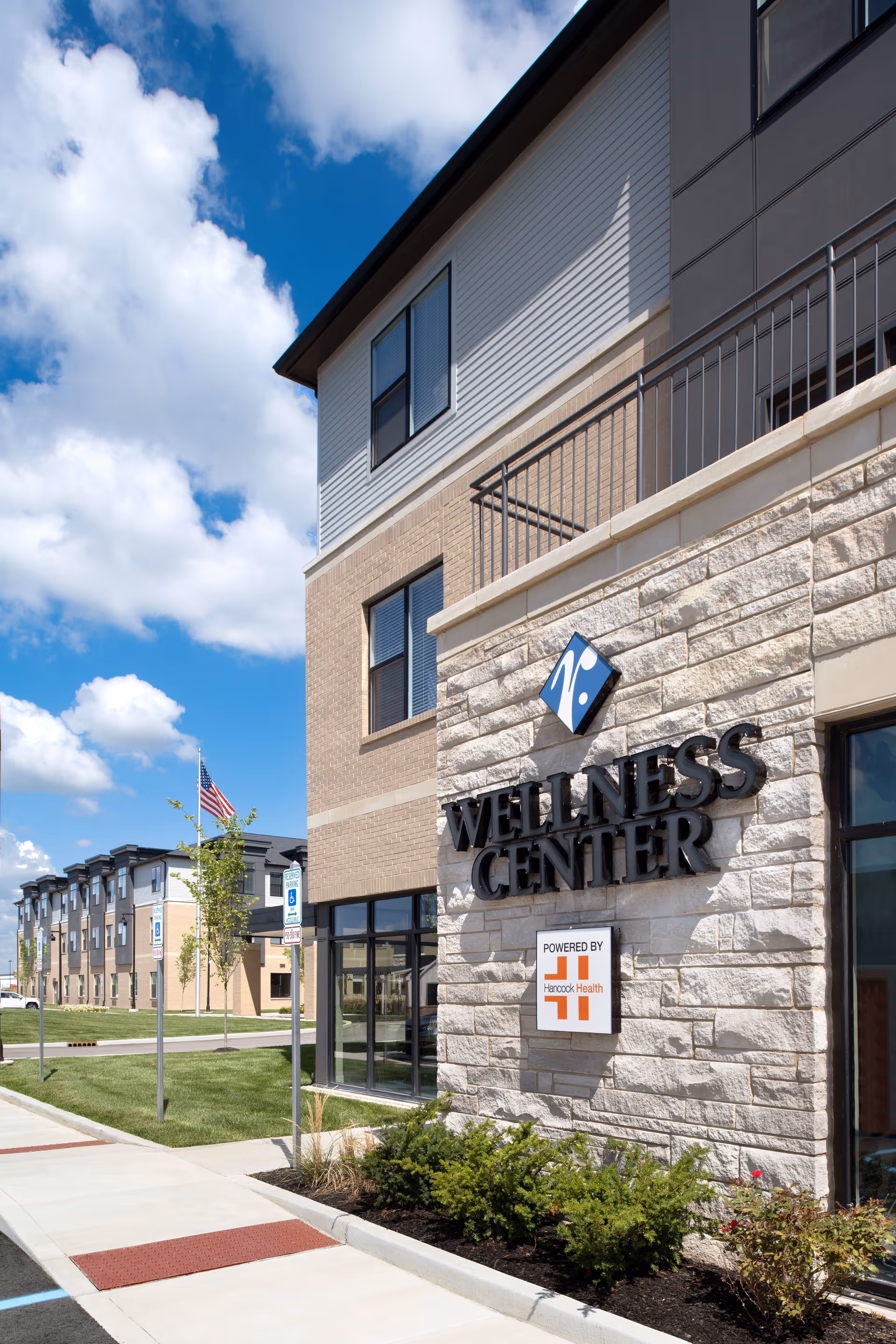 Exterior view of a building with a sign that reads 'Wellness Center' powered by Hancock Health. The building has a stone facade with windows and a sidewalk with grass and small bushes in front. An American flag is visible in the background under a partly cloudy blue sky.