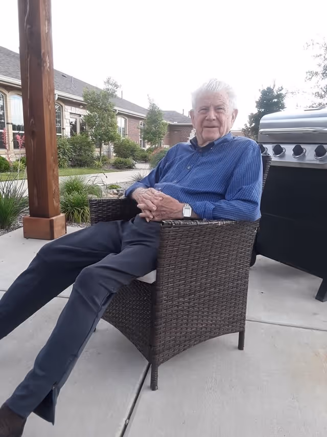 An elderly man wearing a blue shirt and dark pants is sitting and relaxing on a wicker chair outdoors on a concrete patio. Behind him, there is a large grill and a wooden post, with a landscaped garden and a brick building with windows in the background.