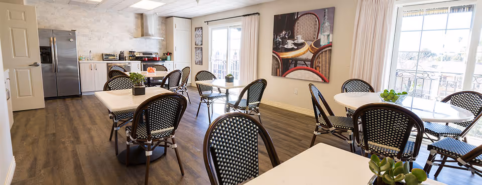 A bright dining area with several tables and black-and-white woven chairs. The room features large windows with white curtains allowing natural light to fill the space. There is a kitchen area in the background with a stainless steel refrigerator, microwave, and cabinets. The floor is wood, and there is a large wall art piece depicting a café scene.
