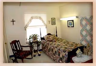 A bedroom in a senior care facility with a single bed, quilt, chair, and plants.