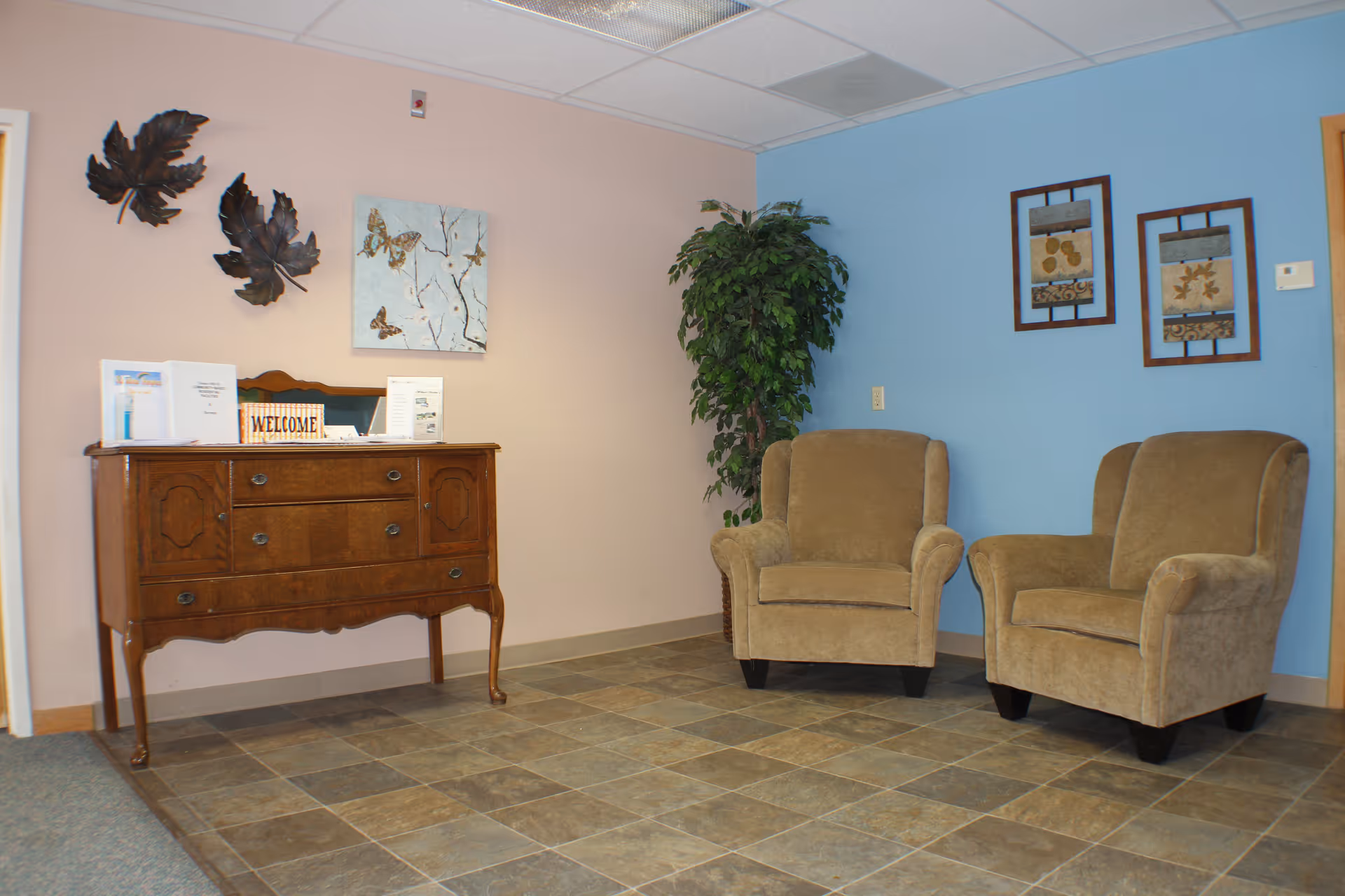 A waiting area with two beige upholstered armchairs against a blue wall. To the left is a wooden sideboard with a welcome sign and informational pamphlets. The walls are decorated with framed artwork and metal leaf sculptures. The floor is tiled with a mix of brown and gray tiles.