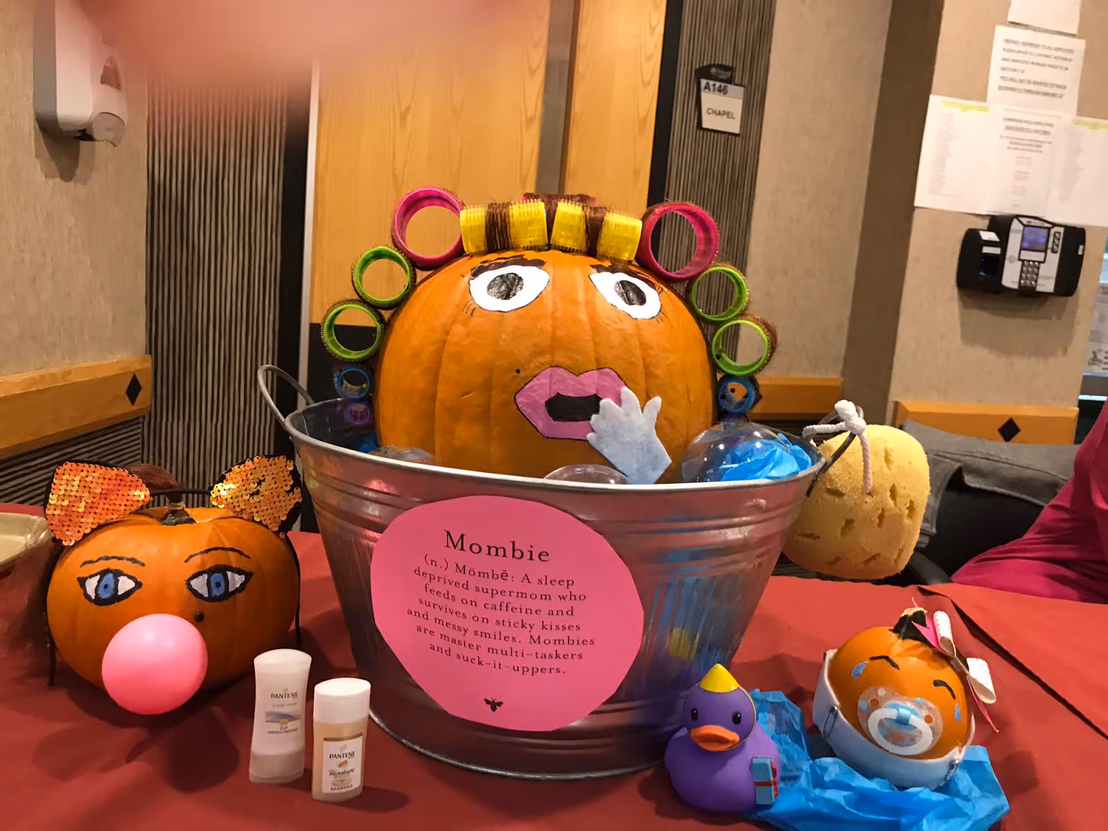 Decorated pumpkins and toys arranged on a table, including a large pumpkin with hair rollers in a metal tub labeled "Mombie".