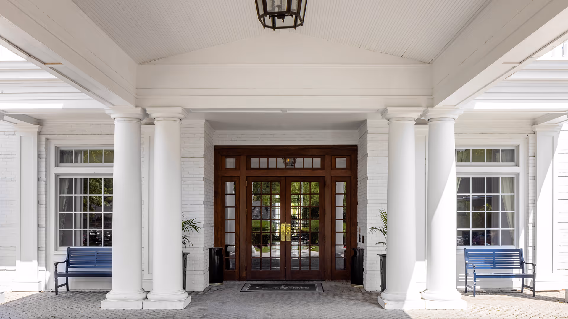Entrance of a senior living facility with large white columns, two blue benches on either side, and double wooden doors with glass panels in the center.