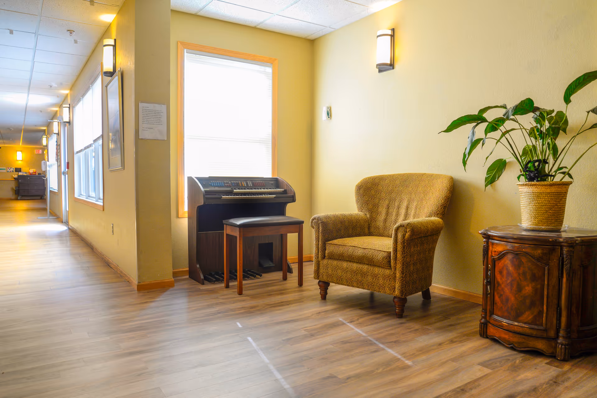 A cozy corner in a senior living facility hallway featuring a patterned armchair, a small wooden organ with a bench, and a wooden side table with a potted plant. The walls are painted light yellow, and the floor is wood laminate. Wall-mounted lights illuminate the space, and windows with blinds let in natural light.