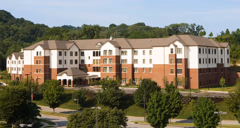 Front exterior of a multi-story brick-and-white senior living building set among trees and landscaped grounds.