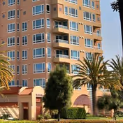 Exterior view of a multi-story residential building with balconies, surrounded by palm trees and greenery under a clear sky.