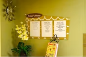 A wall with a decorative clock on the left, a bulletin board displaying today's menu with three sections, and a small stand with a sign featuring colorful text. There are also some green plants at the bottom left corner.