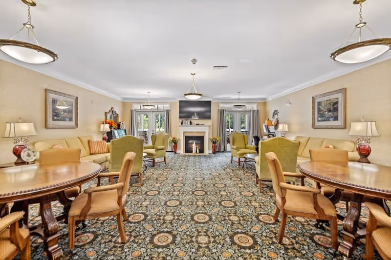 A spacious, well-lit living room with patterned carpet, multiple round wooden tables with chairs, beige sofas, green armchairs, table lamps, framed paintings on the walls, and a fireplace with a mounted flat-screen TV above it. Large windows with curtains allow natural light to enter the room.