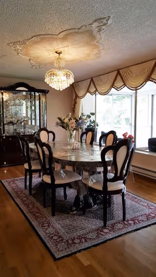 A dining room with a rectangular marble table surrounded by six black and white cushioned chairs. A crystal chandelier hangs from an ornate ceiling medallion above the table. A glass-front china cabinet filled with glassware is against the wall. The room has large windows with decorative valances, and a floral centerpiece is on the table. The floor is hardwood with a patterned area rug under the table.