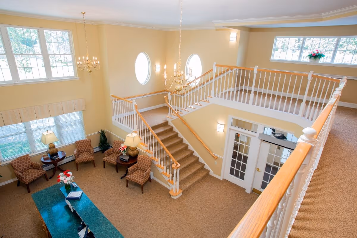 Bright two-story lobby with a carpeted staircase, seating area, chandeliers and large windows.