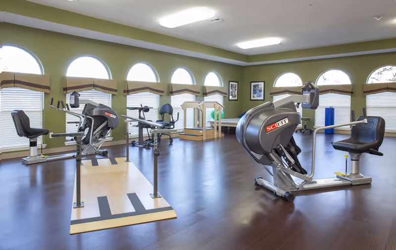 Bright exercise and therapy room with recumbent exercise machines, wooden floor, and arched windows.