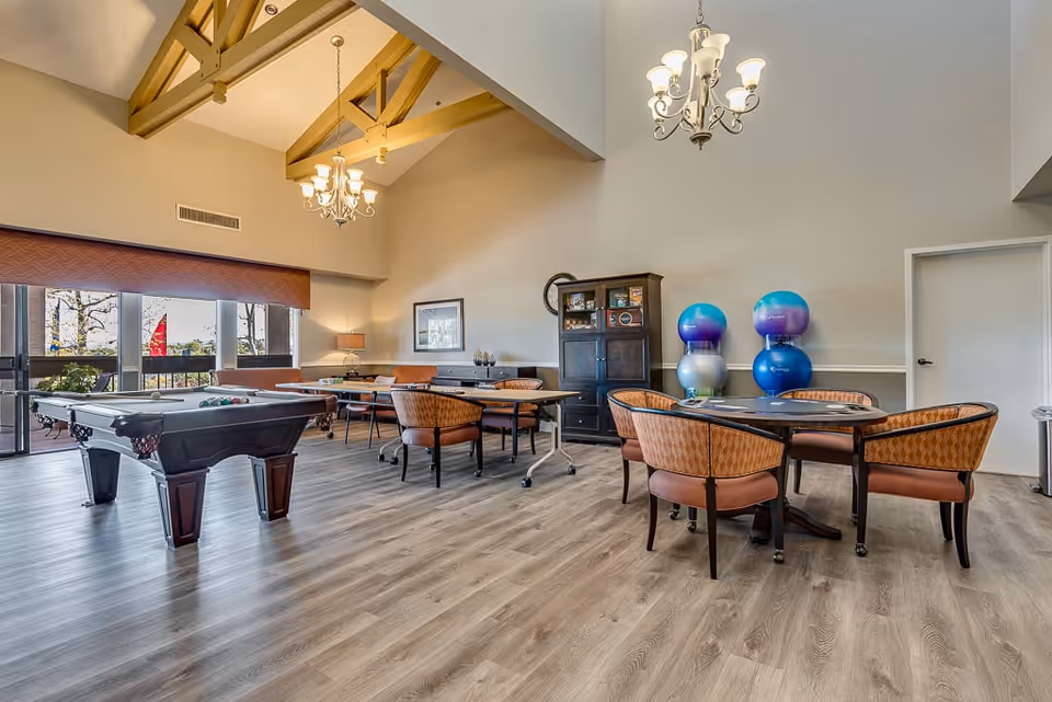 A spacious senior living common area with a pool table, several tables and chairs, exercise balls stacked in the corner, large windows letting in natural light, and chandeliers hanging from a high ceiling with exposed wooden beams.