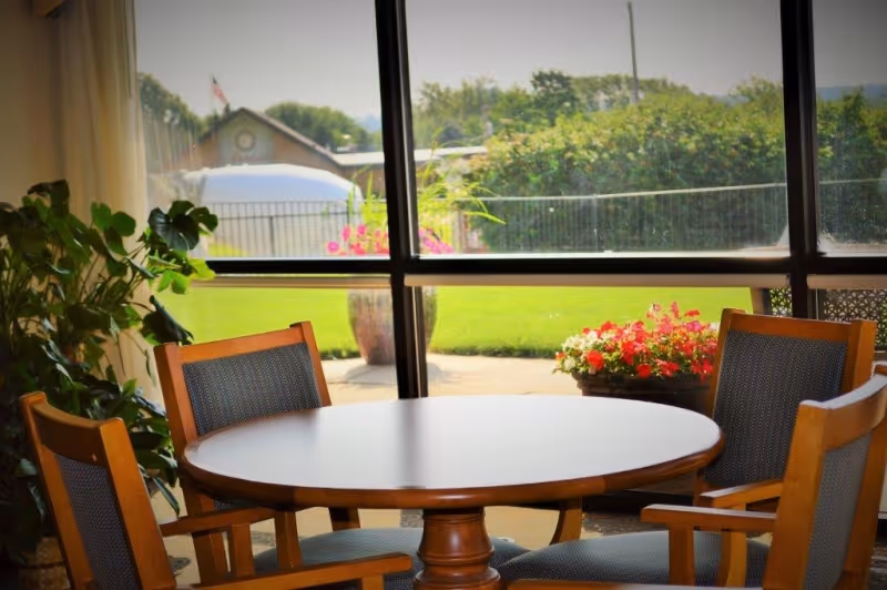 Round wooden dining table with four chairs in front of a large window overlooking a lawn and potted flowers.