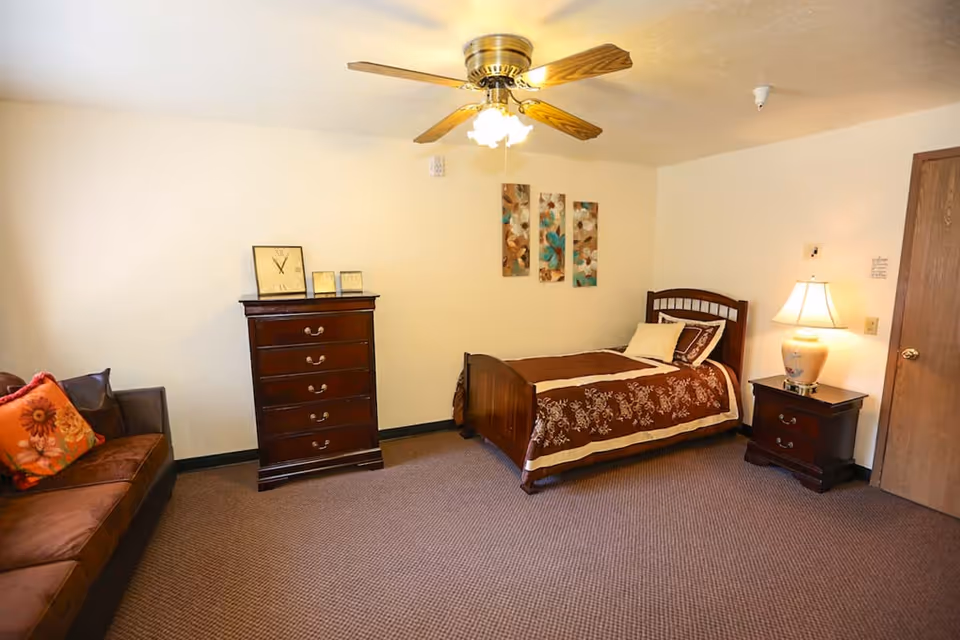 Furnished assisted-living bedroom with a single bed, nightstand and lamp, dresser, ceiling fan, and a sofa.