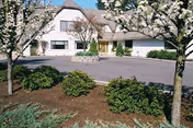 Front entrance of a two-story residential care building with a driveway, landscaped shrubs, and flowering trees.