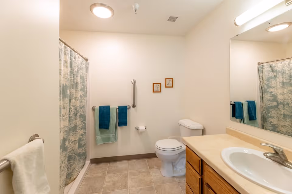 A clean bathroom with beige tiled floor, a white toilet, a wooden vanity with a sink and faucet, a large mirror above the sink, a shower with a patterned curtain, and several towels hanging on towel racks. Two small framed pictures hang on the wall above the toilet.