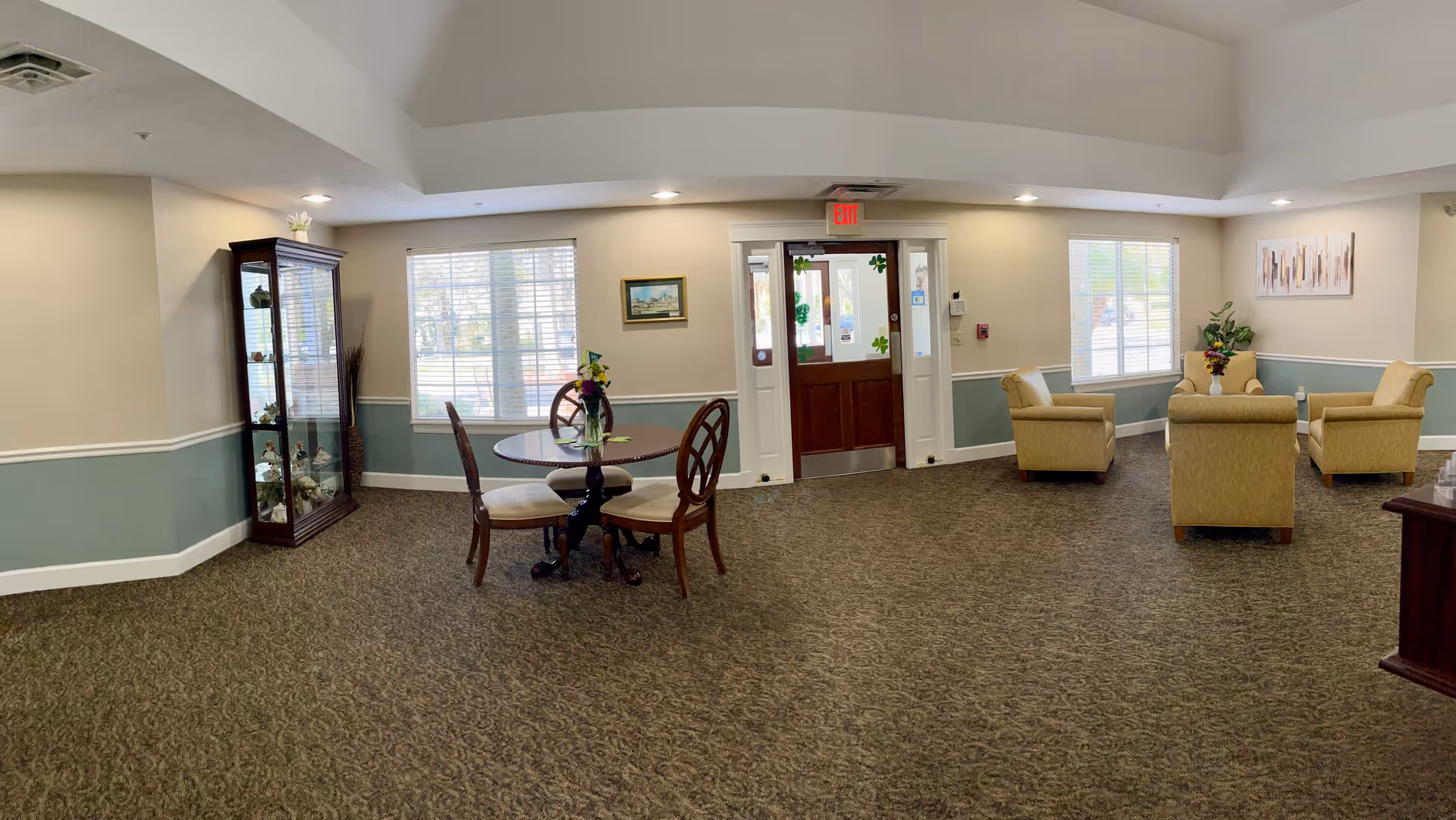 A senior living facility common area with a round wooden table and four chairs in the center, a glass display cabinet with decorative items on the left, and a seating area with four beige armchairs arranged around a small table with a flower vase on the right. There are two large windows with blinds and a wooden exit door with glass panels in the background. The walls are painted beige and light blue with white trim, and the floor is carpeted with a patterned design.