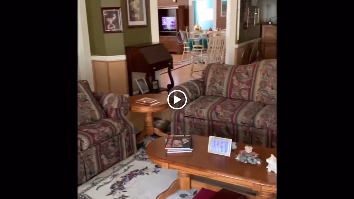 A cozy living room with patterned upholstered armchairs and a sofa arranged around a wooden coffee table. The coffee table holds books, a framed photo, and small decorative figurines. In the background, there is a wooden writing desk, framed pictures on the walls, and a dining area with a table and chairs. The room has a warm, homey atmosphere with green and beige walls and wooden flooring.