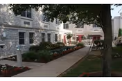 Outdoor courtyard area of a senior living facility with a paved walkway, flower beds, and trees. The building has multiple windows and light-colored stone exterior walls. There are benches and a swing set visible along the walkway.