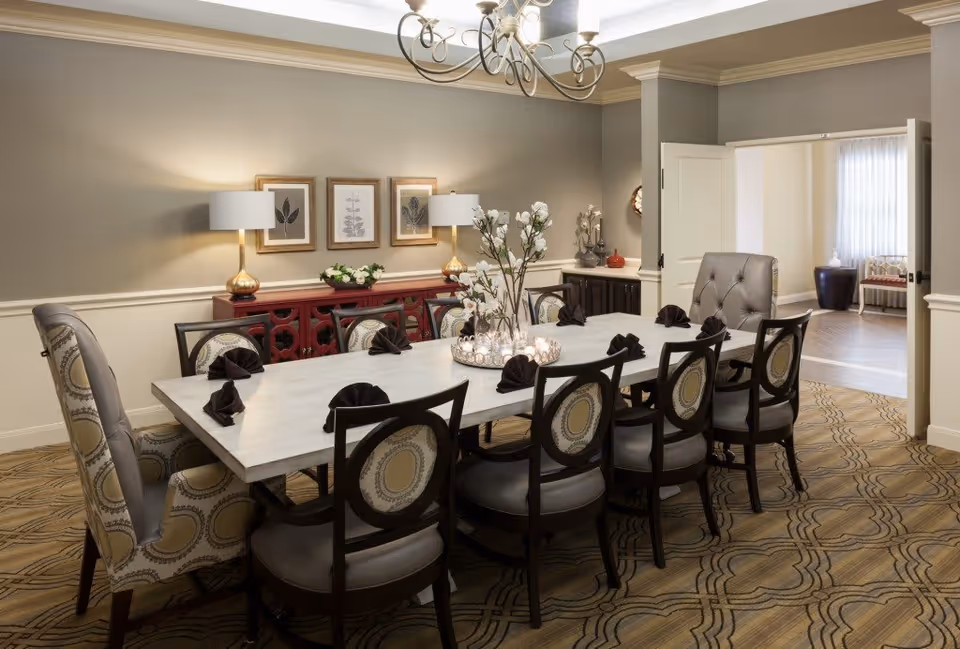 A formal dining room with a long rectangular table surrounded by ten chairs, including two armchairs at the ends. The table is decorated with black folded napkins and a centerpiece with flowers and candles. The room has beige walls with white trim, a patterned carpet, two table lamps on a red sideboard, and three framed botanical prints on the wall. A chandelier hangs from the ceiling, and an open doorway leads to another room with a bench and window.