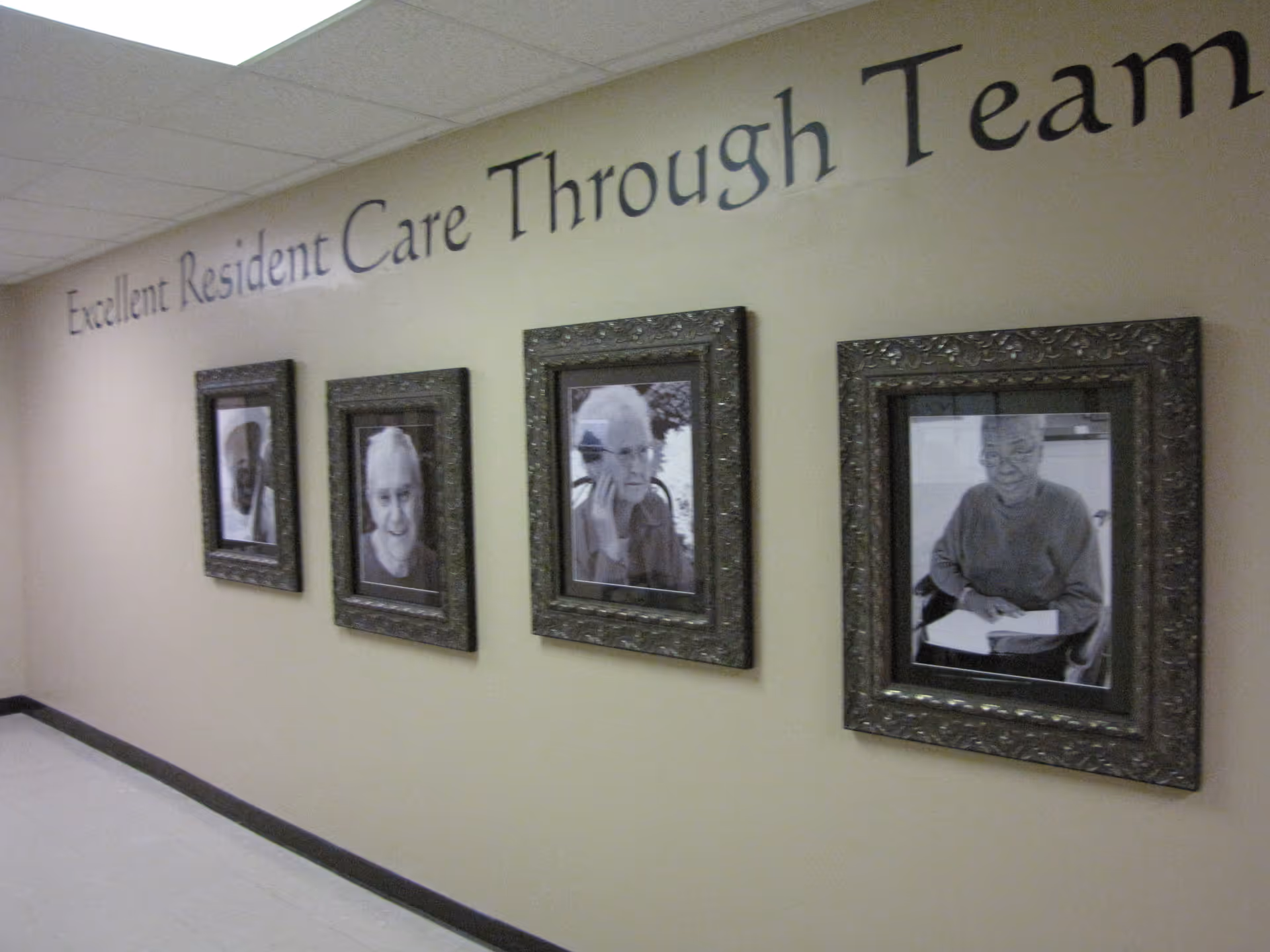 A beige hallway wall with four framed black and white portraits of elderly individuals. Above the frames, the text on the wall reads 'Excellent Resident Care Through Team'.