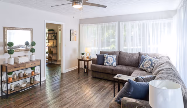 A bright and spacious living room with large windows covered by sheer white curtains allowing natural light to fill the space. The room features a large L-shaped gray sofa adorned with patterned and solid blue throw pillows. There is a wooden side table with a lamp and a small plant, and a console table with decorative items and a mirror above it. The floor is dark wood, and a ceiling fan is mounted on the white ceiling.