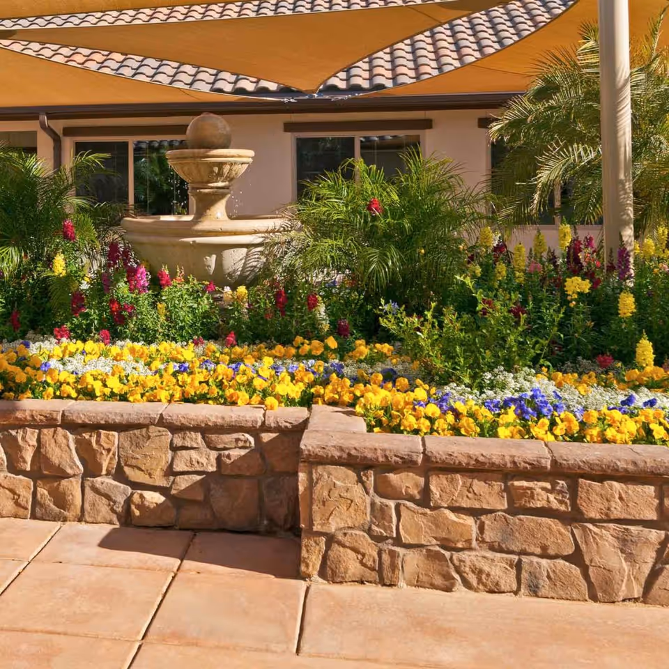 A vibrant outdoor garden area with a stone planter filled with colorful flowers including yellow, purple, and red blooms. In the background, there is a beige building with a tiled roof and windows, partially shaded by large tan fabric sunshades. A decorative stone water fountain is centered among green plants and flowers.