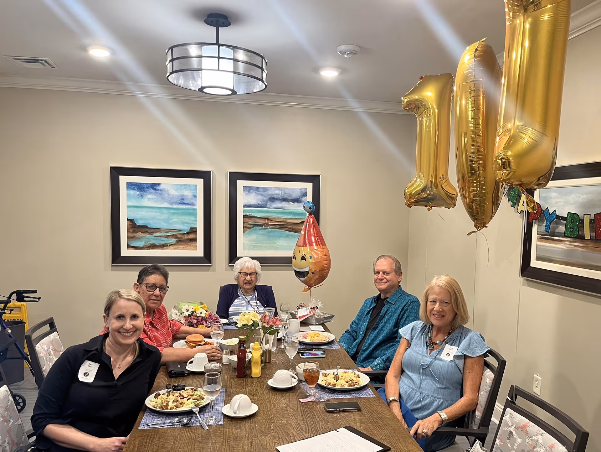 Five people sitting around a dining table in a room decorated with balloons shaped as the number 101 and a happy birthday banner. The table has plates of food, drinks, and flowers. Two framed paintings hang on the wall behind them.