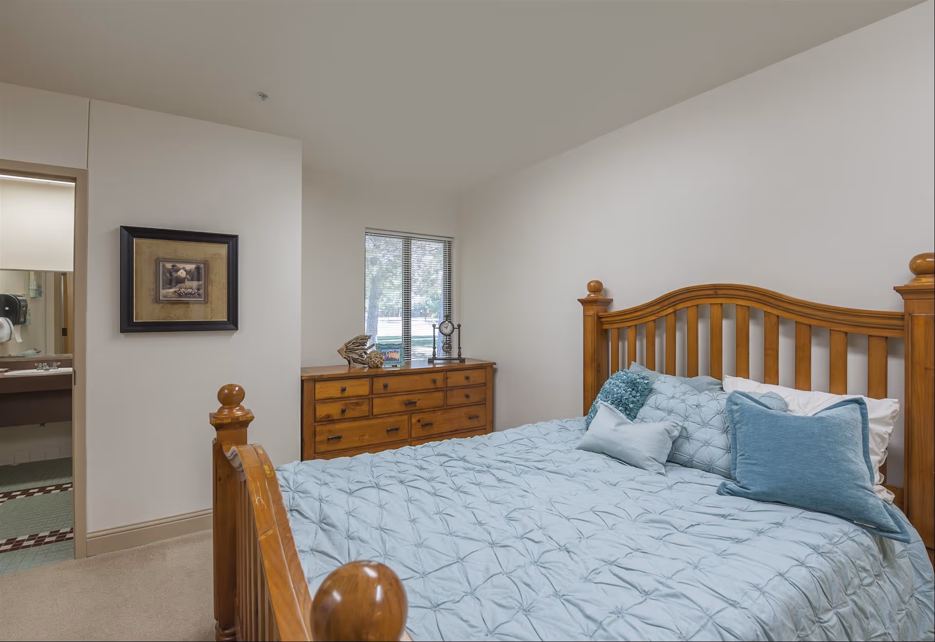 A bedroom in an assisted living facility featuring a wooden bed frame with a light blue comforter and multiple pillows. There is a wooden dresser with decorative items on top, a window with blinds, and a framed picture on the wall. A bathroom is partially visible through an open door.