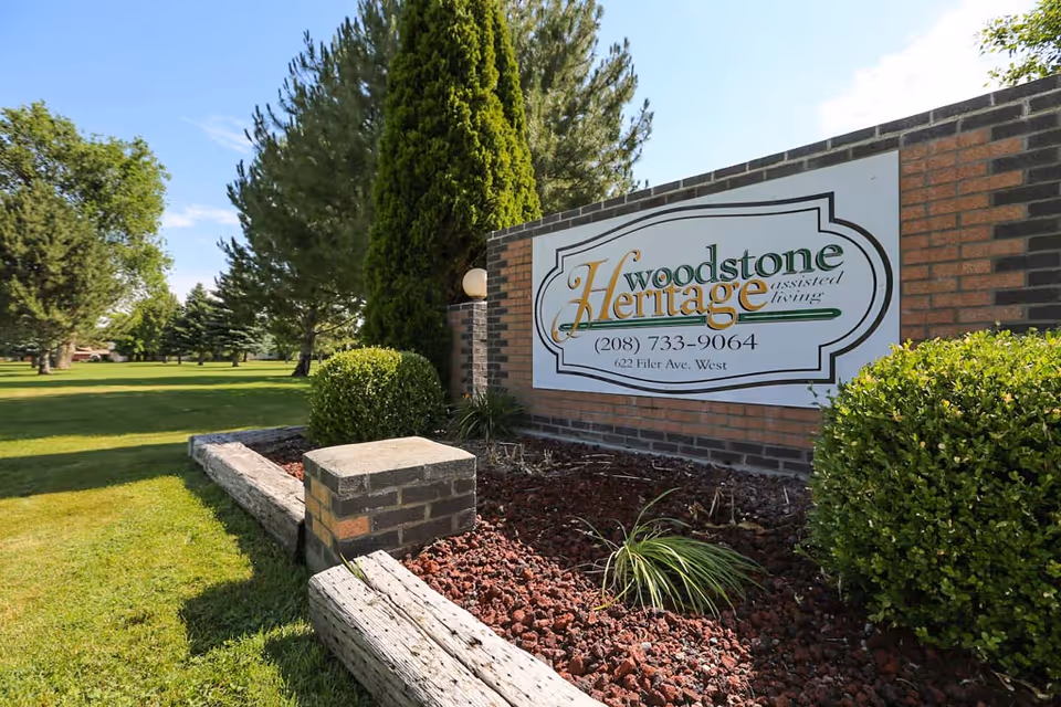 Outdoor view of a landscaped area with green grass, trees, and bushes surrounding a brick sign for Woodstone Heritage Assisted Living, displaying a phone number and address.