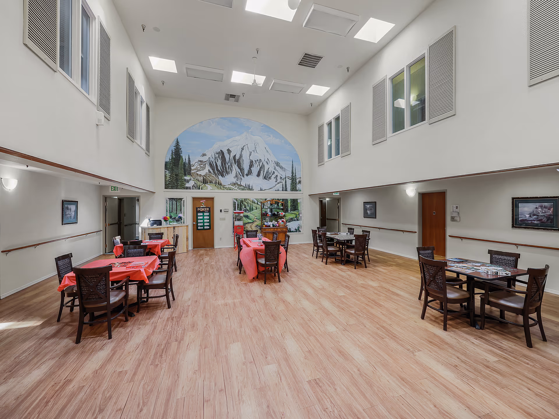 A spacious, well-lit common room with high ceilings and large windows near the ceiling. The room has several tables with chairs arranged around them, some tables covered with red tablecloths. A large mural of a snow-capped mountain and pine trees decorates the far wall. The floor is wooden, and the walls are painted white with handrails along them. There are doors and framed pictures on the walls.