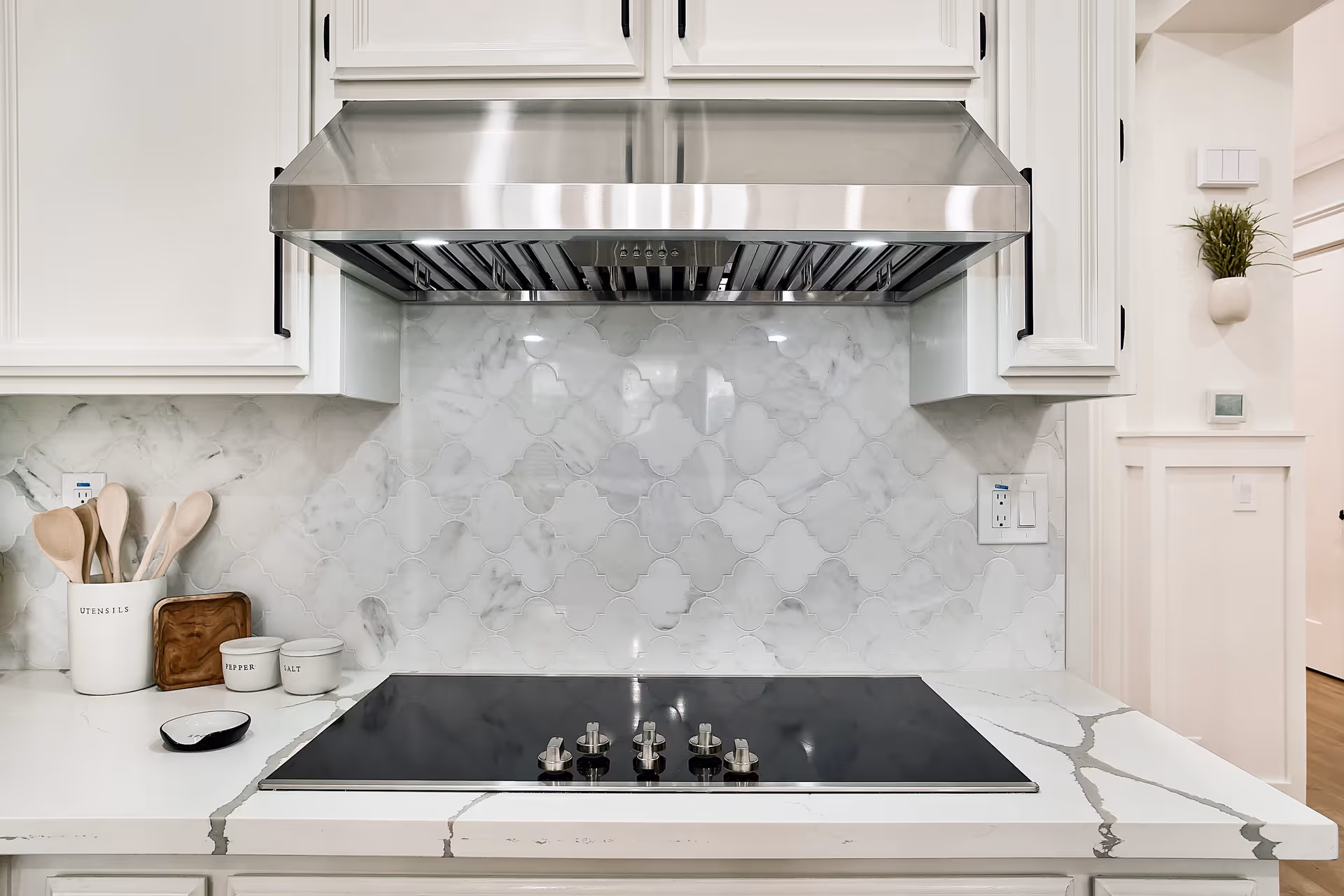Modern kitchen stovetop with five control knobs on a white marble countertop. Above the stovetop is a stainless steel range hood and white cabinetry. The backsplash features a white and gray patterned tile. On the left side of the counter are containers labeled utensils, pepper, and salt, along with wooden cooking spoons. A small plant is mounted on the wall to the right.