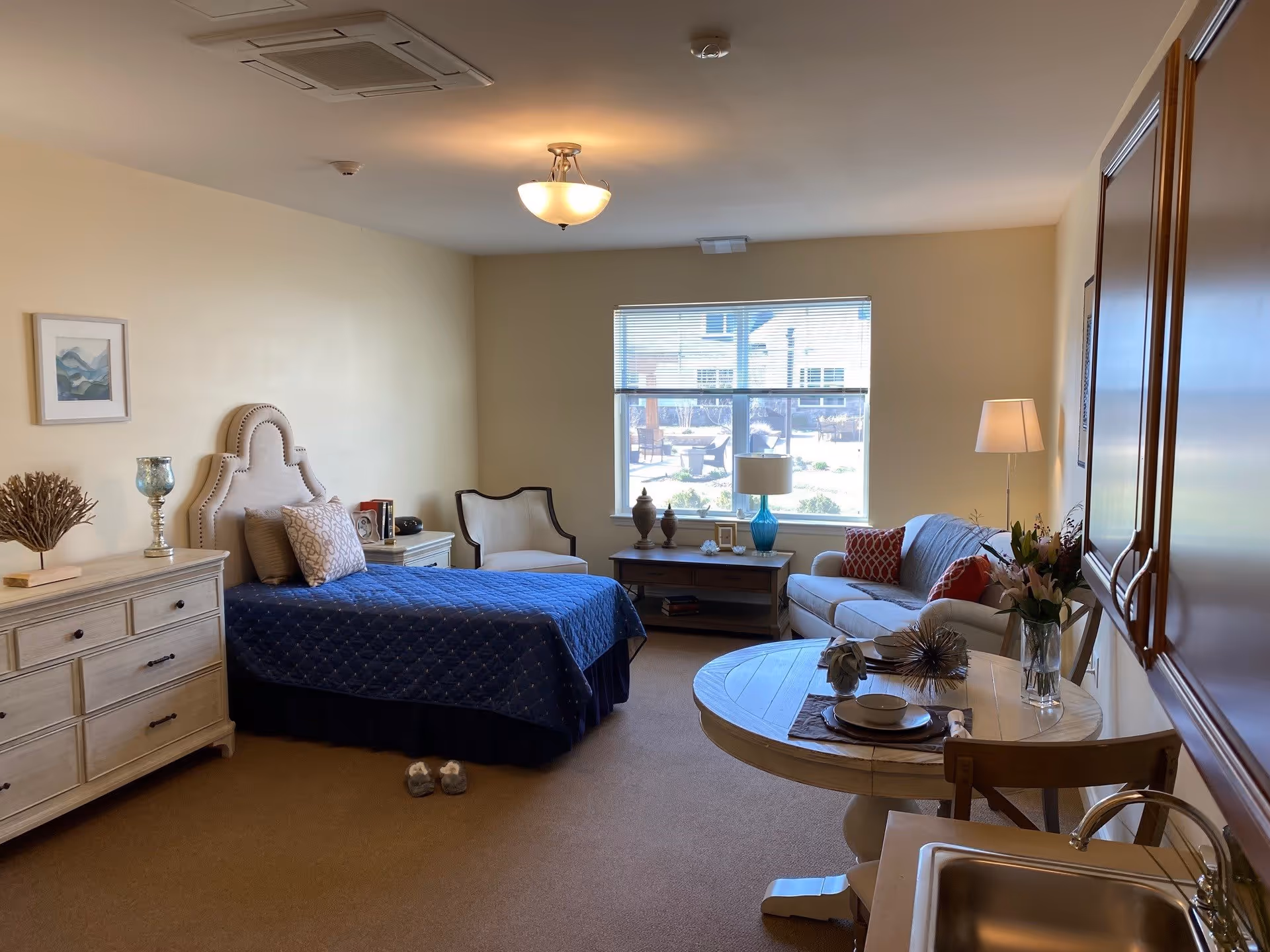 A cozy senior living room with a single bed covered in a blue quilt, a beige armchair, a white sofa with red pillows, a round wooden table set with plates and bowls, a dresser with decorative items, and a window letting in natural light.