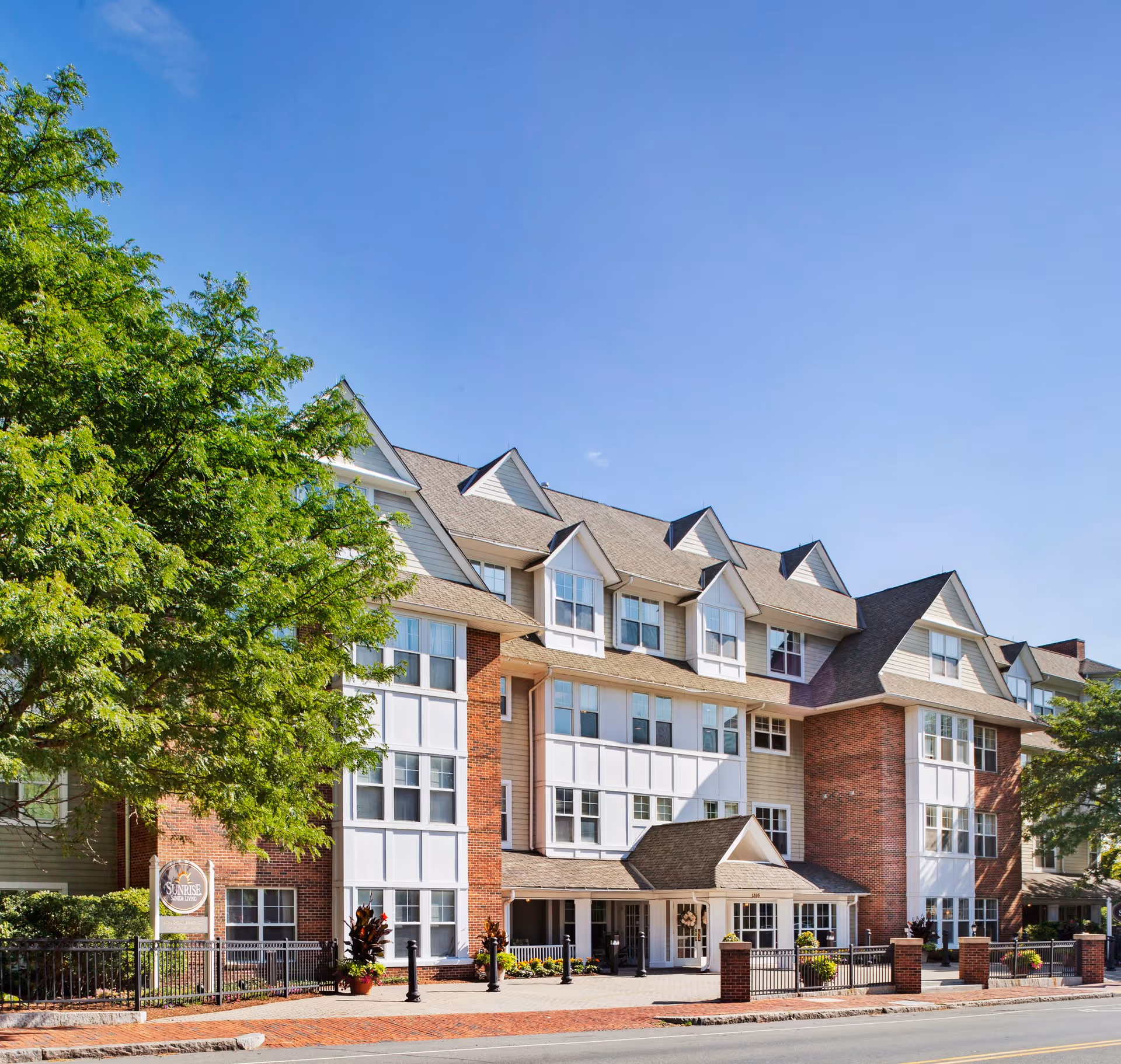 Exterior view of a multi-story senior living facility building with brick and white paneling, multiple windows, and a covered entrance. There are trees and plants around the building, and a clear blue sky above.