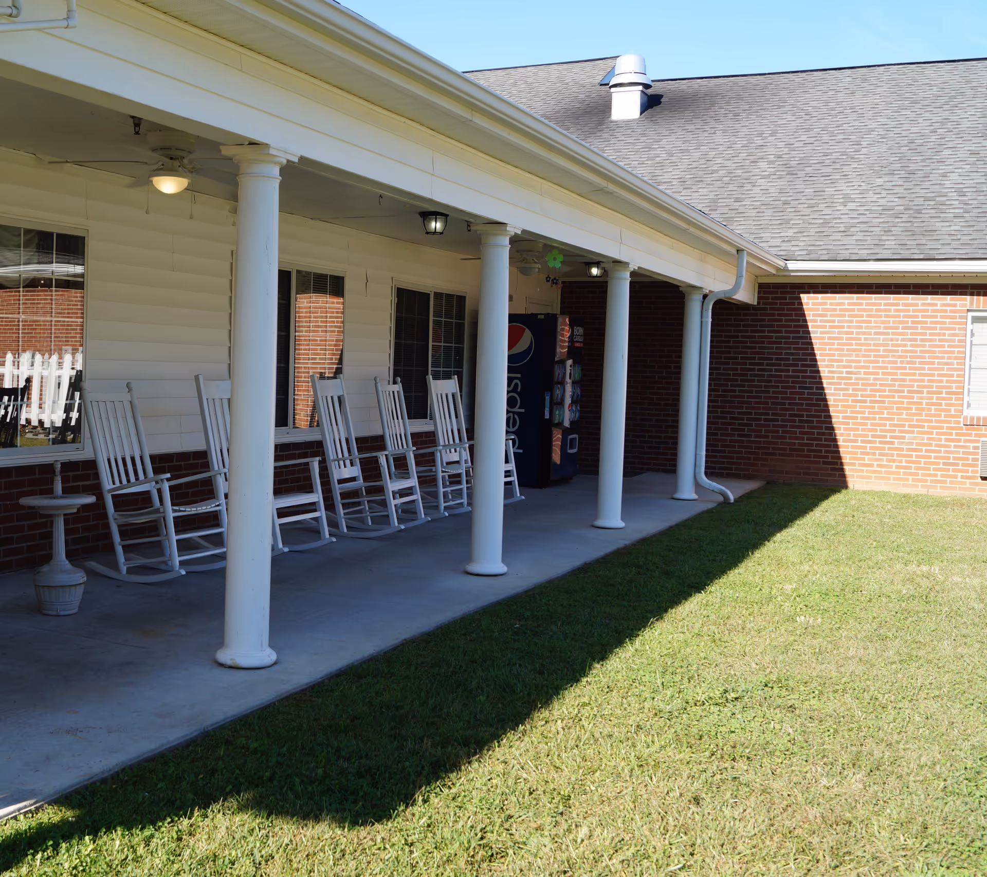 Covered exterior porch with white columns and a row of white rocking chairs next to a brick wall and a vending machine.