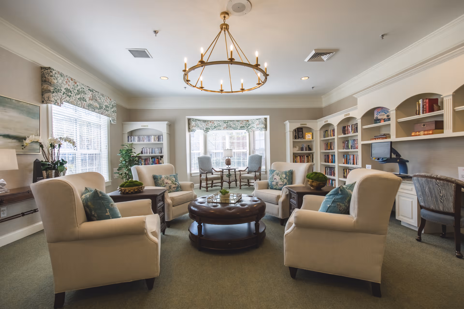 A cozy and well-lit common area with four beige armchairs arranged around a round leather ottoman. The room features built-in white bookshelves filled with books, a computer workstation, and large windows with floral valances letting in natural light. A circular chandelier hangs from the ceiling, and there are decorative plants and cushions adding to the inviting atmosphere.