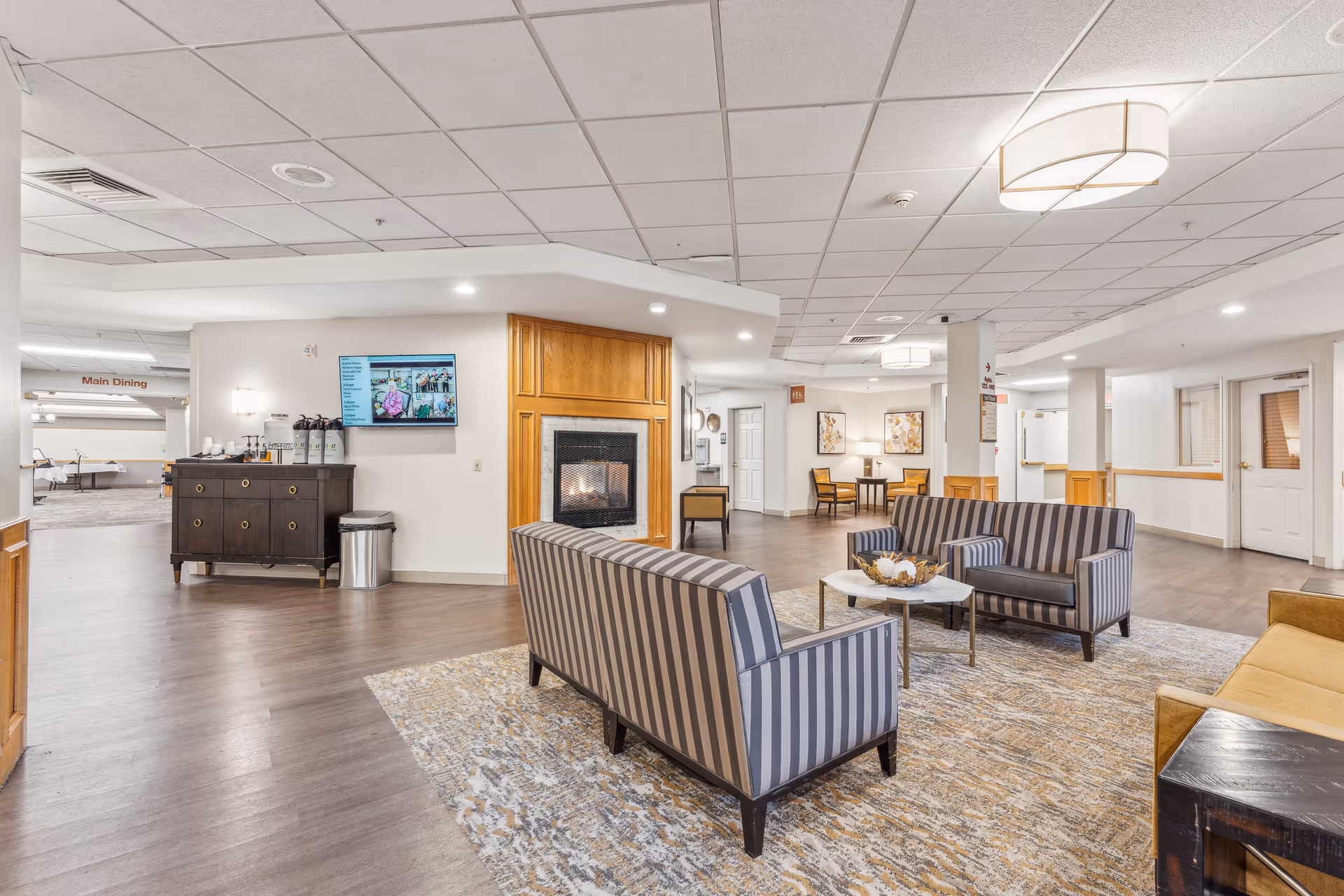Well-lit senior living lounge with striped sofas arranged around a coffee table, a fireplace, and a nearby coffee station.