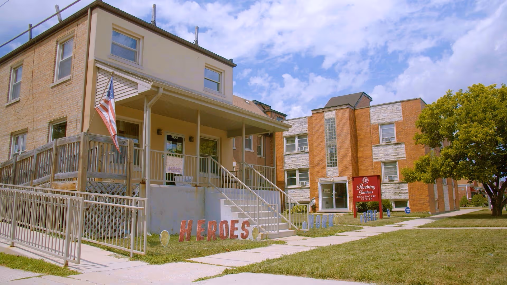 Front of a brick senior care building with a ramp and steps, an American flag, and 'HEROES' painted on a low wall in the foreground.