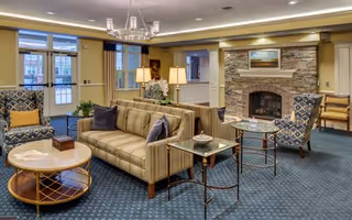A cozy living room area with a striped sofa, two patterned armchairs, a round wooden coffee table, and two glass side tables. The room features a stone fireplace with a painting above it, a chandelier, and large windows letting in natural light.