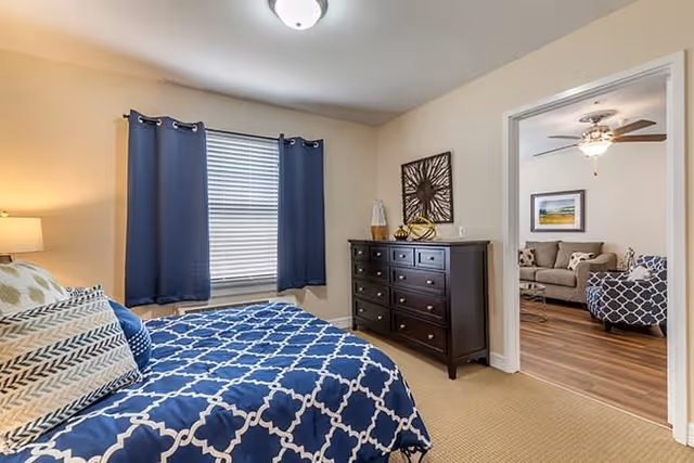 A bedroom with a bed covered in a blue and white patterned comforter and multiple pillows. There is a window with blue curtains, a dark wooden dresser with decorative items on top, and an open doorway leading to a living room with a sofa, armchair, ceiling fan, and framed artwork on the wall.