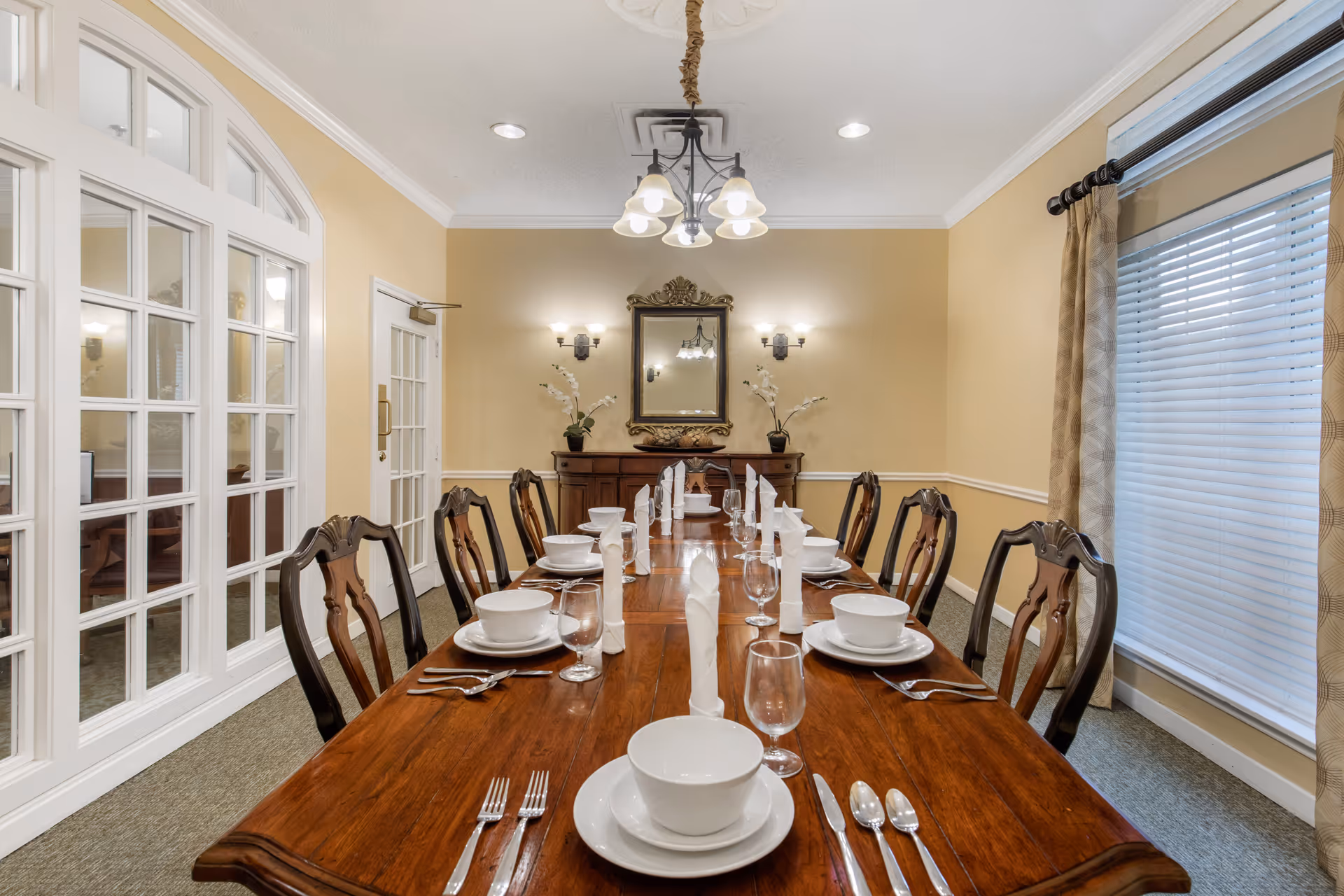 A formal dining room with a long wooden table set for ten people. Each place setting includes a white bowl, plate, glass, and silverware with a folded white napkin. The room has beige walls, a large window with blinds and curtains on the right, and a wooden sideboard with a decorative mirror and two potted plants on the back wall. A chandelier with five lights hangs above the table, and there are wall sconces on either side of the mirror. French doors and glass-paneled windows are on the left side of the room.