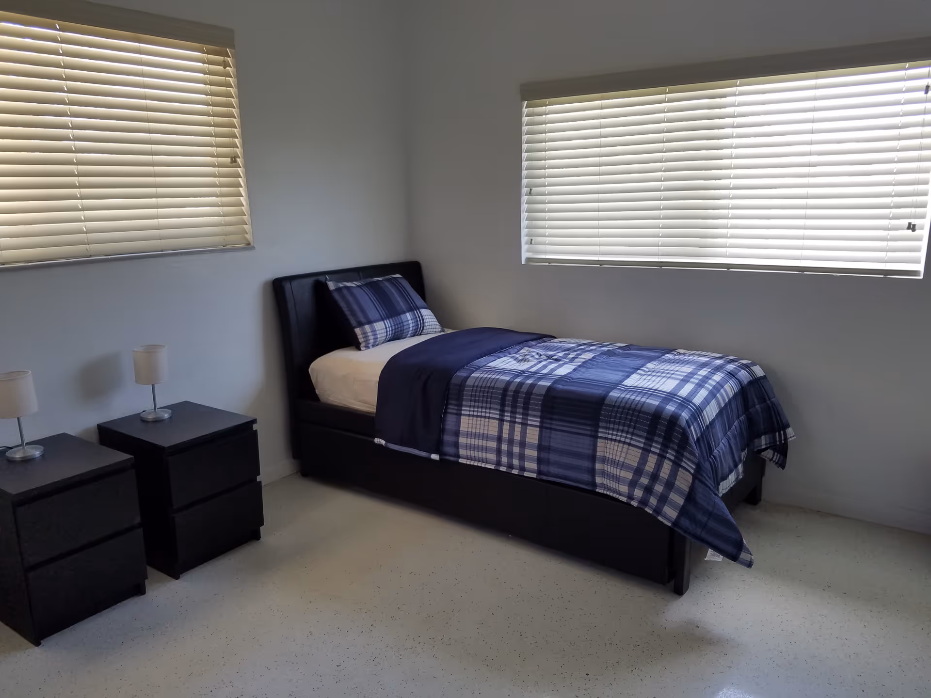 Minimal bedroom with a single bed dressed in blue plaid bedding, two nightstands with lamps, and horizontal blinds on the windows.