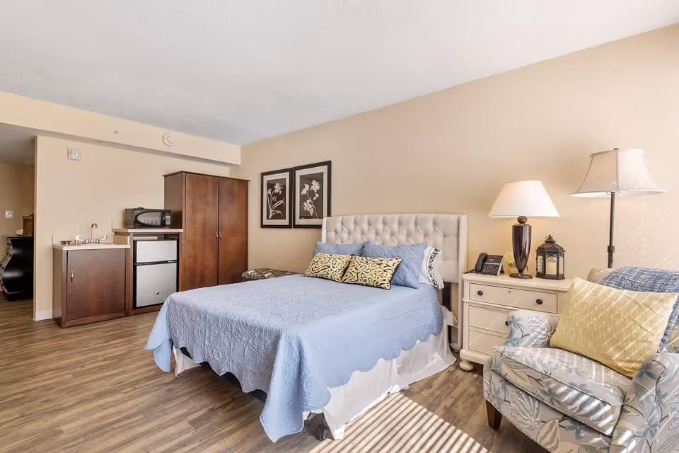 A senior living facility bedroom featuring a bed with a light blue quilt and decorative pillows, a nightstand with a lamp, phone, and lantern, a floral upholstered armchair with a yellow pillow, and a small kitchenette area with a sink, microwave, mini fridge, and wooden cabinet. The room has light beige walls and wood flooring.