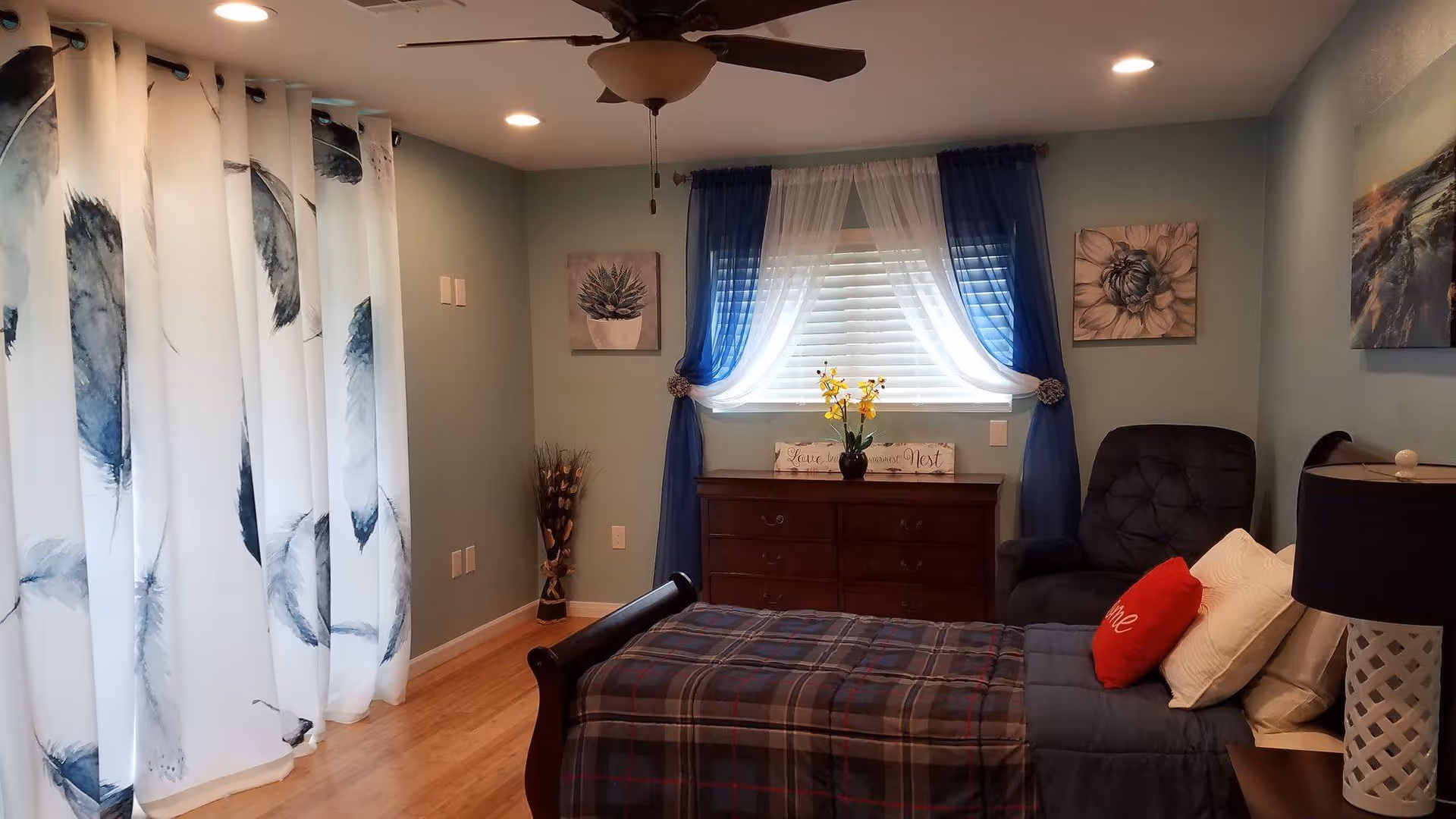 A cozy bedroom with a single bed covered in a plaid blanket and pillows, including a red one with the word 'Home'. There is a dark cushioned armchair next to the bed, a wooden dresser under a window with blue and white curtains, and decorative wall art featuring plants and flowers. The room has light blue walls, wooden flooring, a ceiling fan with lights, and a table lamp with a dark shade on a bedside table.