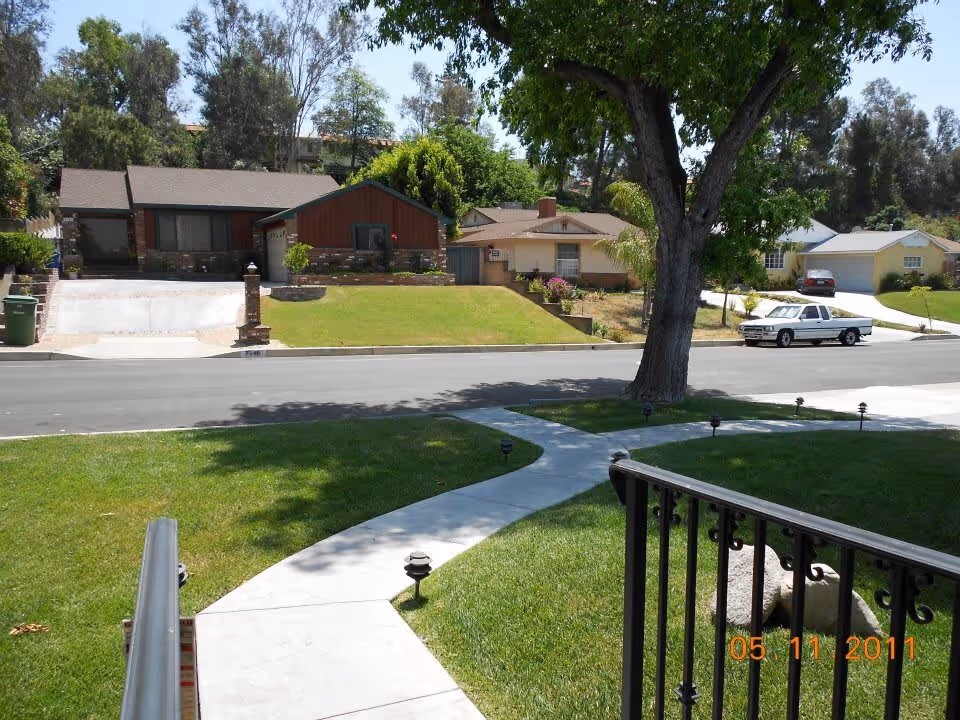 View of a suburban neighborhood street with houses, green lawns, a large tree, and a curved concrete sidewalk leading from a porch with a black metal railing.