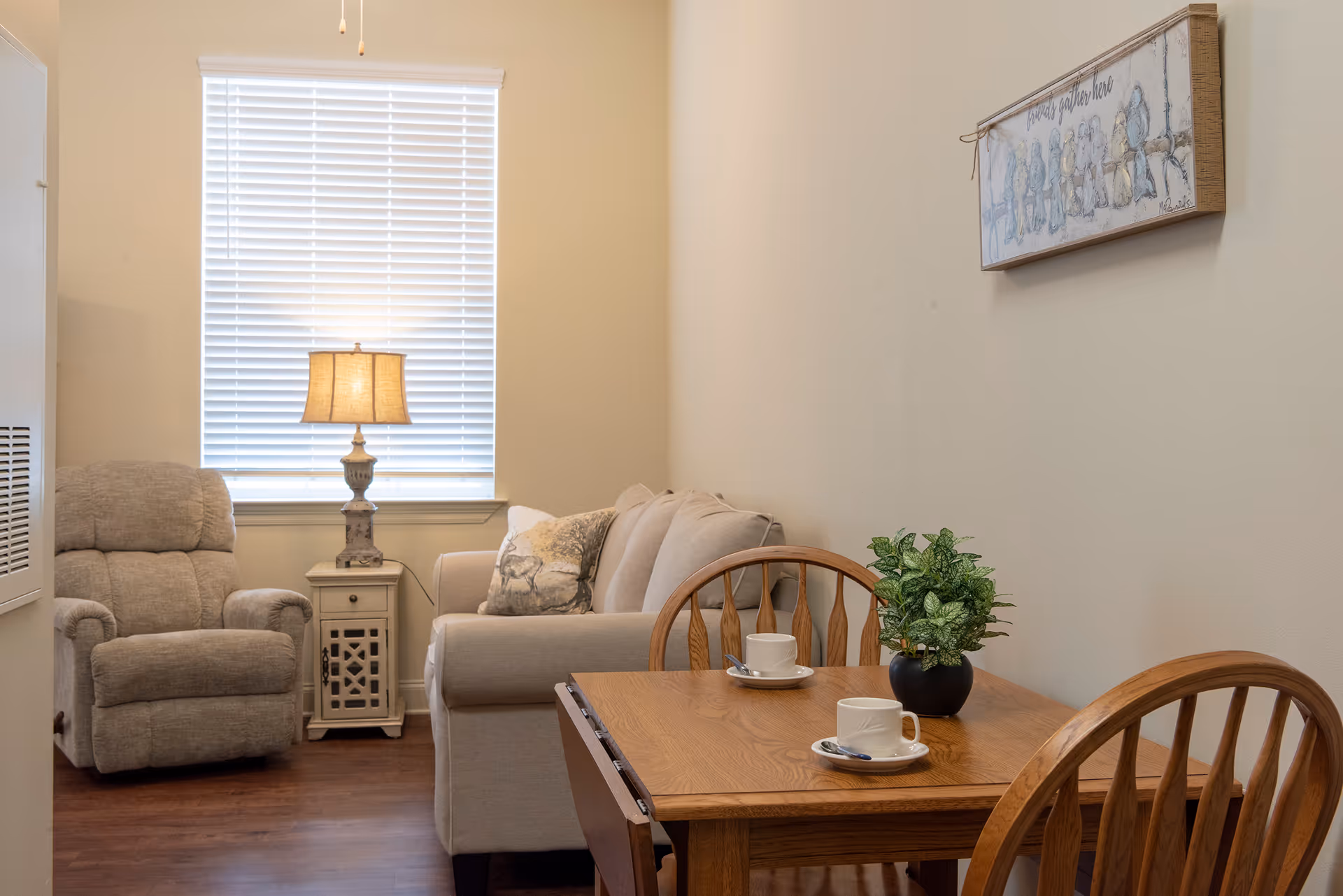 Cozy living room with a recliner and sofa by a window, a side table lamp, and a small wooden dining table with two cups and a potted plant.
