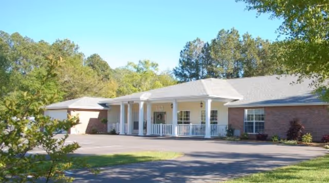 Front exterior view of a single-story brick assisted living facility with a white porch supported by columns, surrounded by trees and greenery under a clear blue sky.