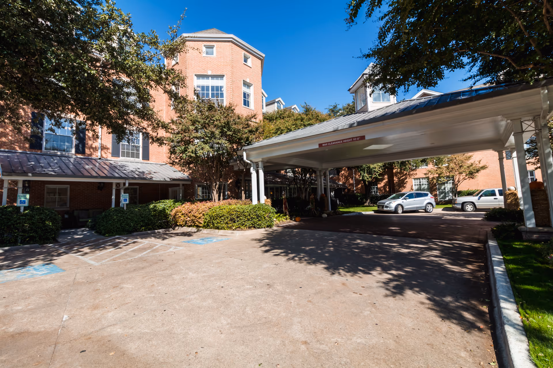 Exterior view of Caruth Haven Court showing a brick building with multiple windows, a covered entrance with a sign indicating max clearance height, several parked cars, trees, and landscaped bushes under a clear blue sky.