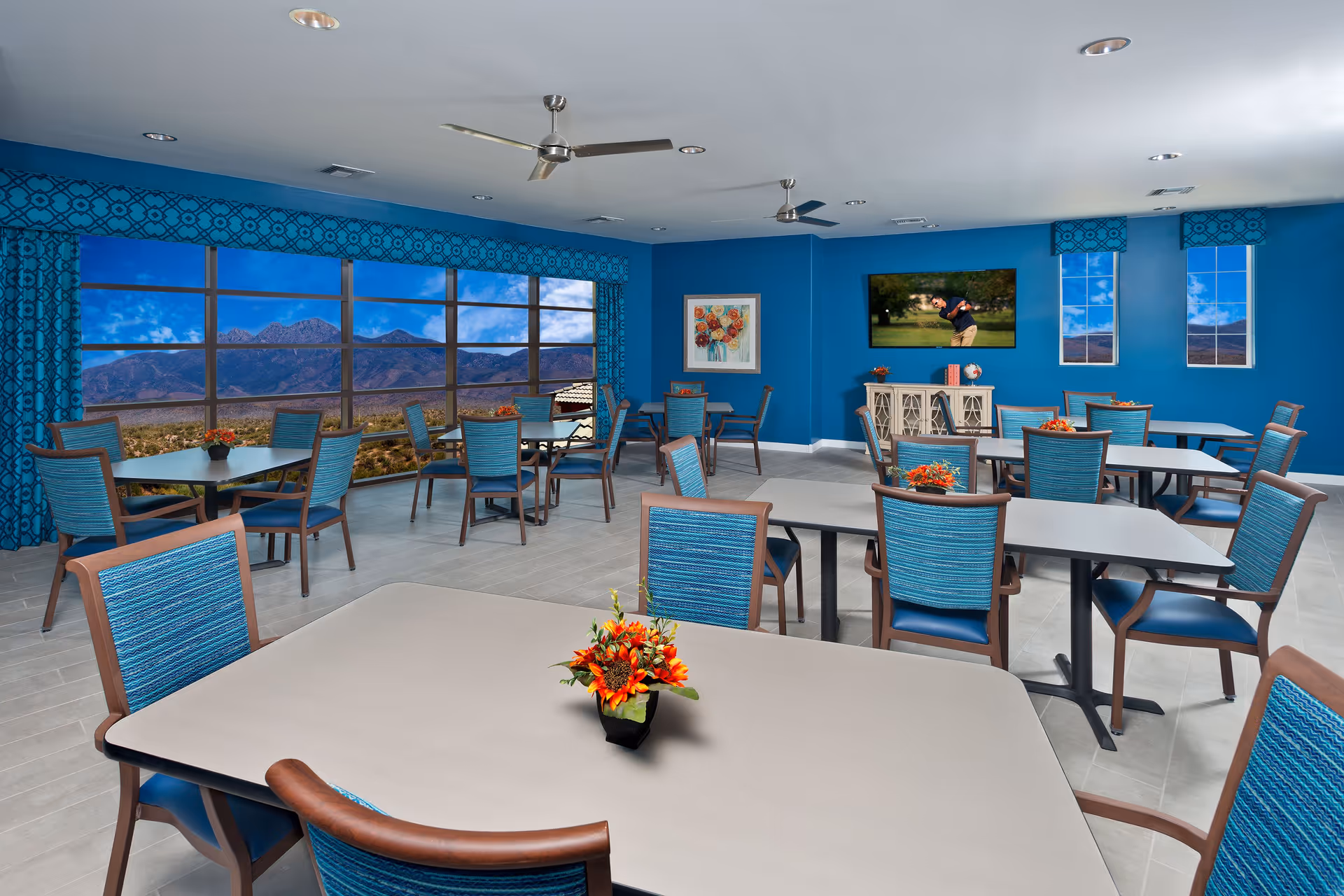 A spacious dining room with multiple tables and blue cushioned chairs. The walls are painted blue and decorated with a floral painting and a flat-screen TV showing a golfer. Large windows with blue patterned curtains offer a scenic view of mountains and a clear sky. Small flower arrangements are placed on each table.